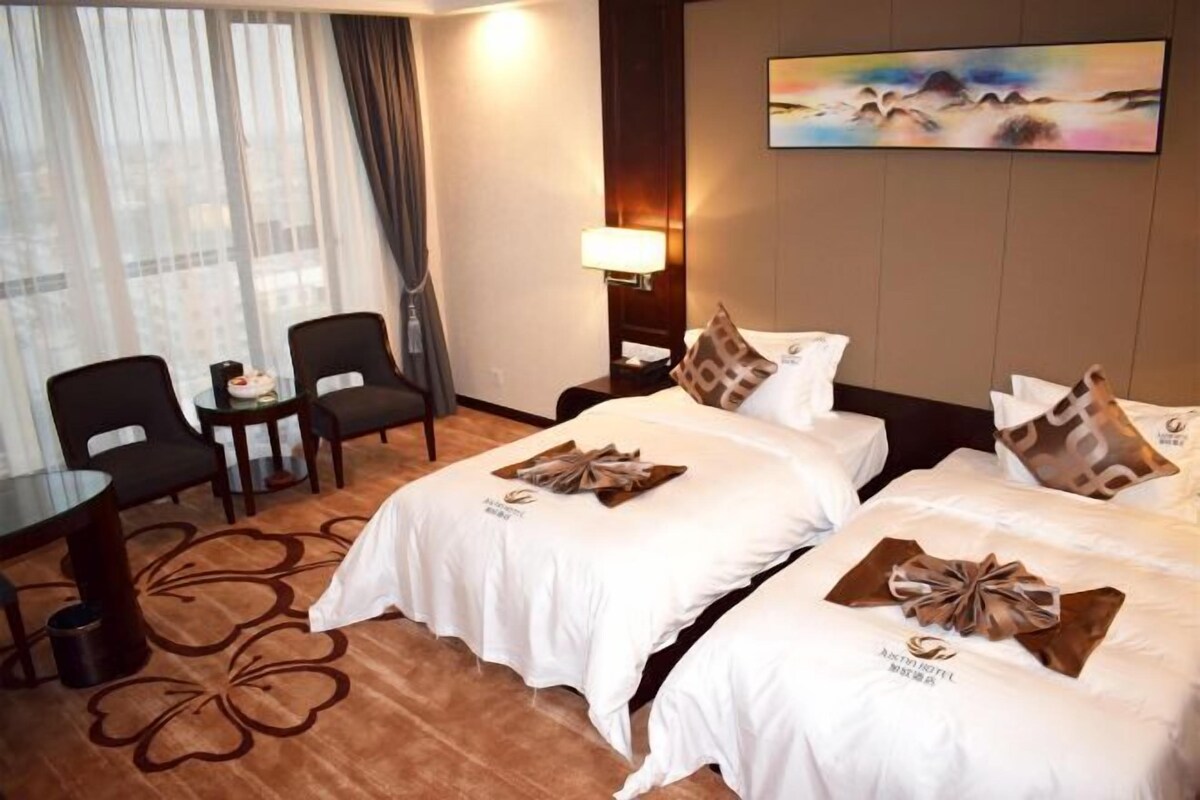 A hotel room with two beds, a desk, chairs, a lamp, and a painting on the wall.