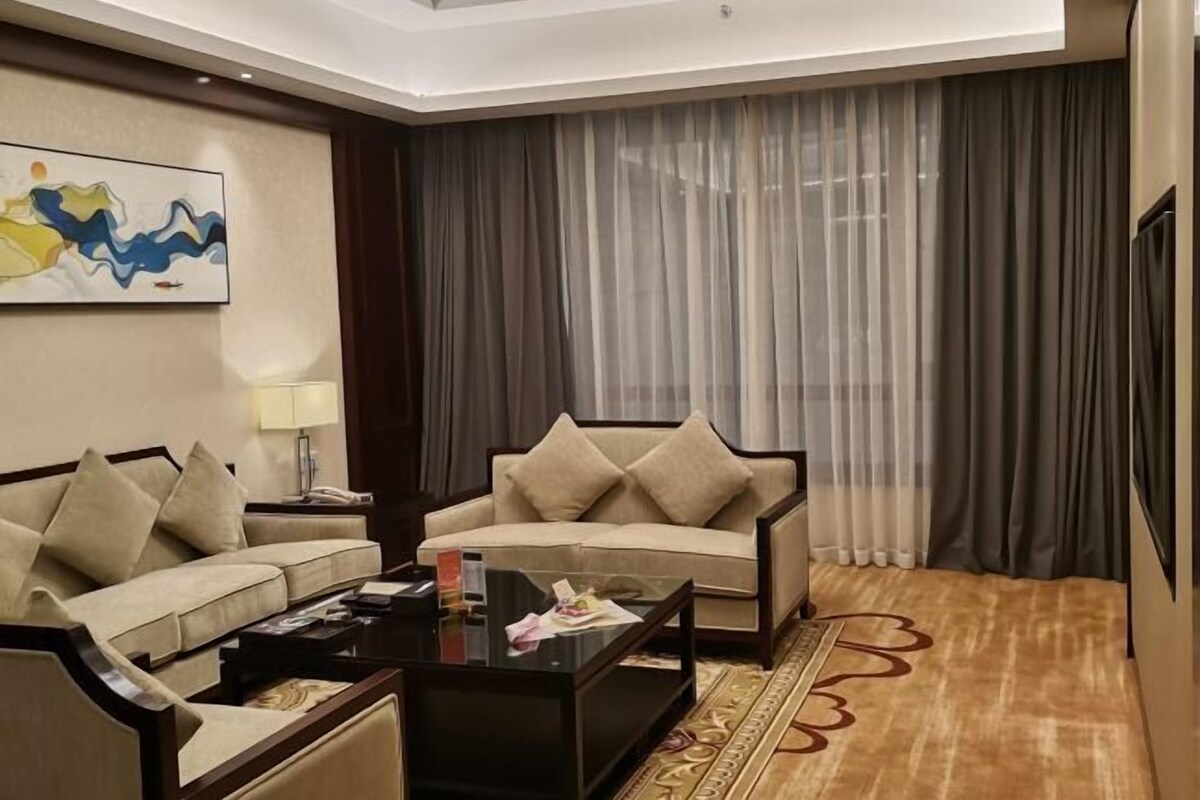 A hotel room with a beige sofa, a smaller armchair, a coffee table, and a large window with sheer curtains.