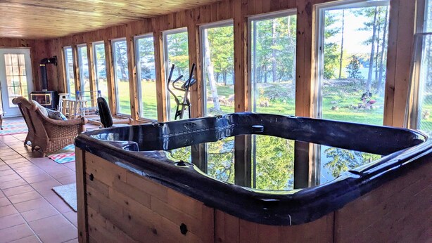 A wooden cabin interior with a hot tub, large windows, and a view of the outdoors. - Serene, Spacious Waterfront Cottage in Kawartha Lakes w. Hot Tub. Pet-Friendly! (Trent Lakes)