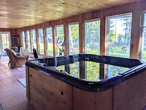 A wooden cabin interior with a hot tub, large windows, and a view of the outdoors. - Serene, Spacious Waterfront Cottage in Kawartha Lakes w. Hot Tub. Pet-Friendly! (Trent Lakes)