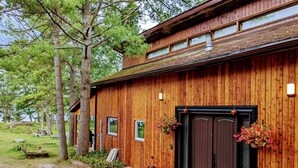 A wooden cabin with a large front door, two windows, and a small porch area with a bench and a potted plant. - Serene, Spacious Waterfront Cottage in Kawartha Lakes w. Hot Tub. Pet-Friendly! (Trent Lakes)