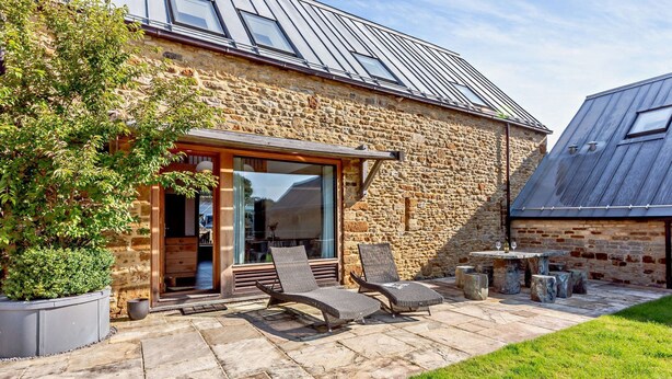 A stone building with a large glass door, outdoor seating, and a metal roof. - Beautifully presented Cotswold holiday cottage with a hot tub - Walnut Barn (Great Tew)