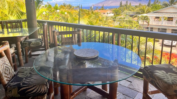 A glass-top table with wooden chairs on a balcony with a view of a mountain and tropical vegetation. - Mango Suite (Kihei)