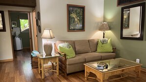 A living room with a sofa, two side tables, a glass coffee table, and a painting on the wall. - Mango Suite (Kihei)
