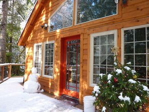Una baita di legno con una porta rossa, ampie finestre e un portico coperto di neve.