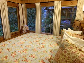 Una camera da letto con una coperta a motivi floreali, ampie finestre con tende e vista su alberi.