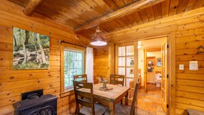 Interior de una cabaña de madera con una mesa de comedor, sillas, un horno de leña y un cuadro en la pared.