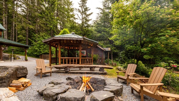 Un belvédère en bois avec un foyer au centre, entouré de fauteuils Adirondack, situé dans une zone boisée.