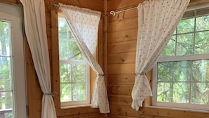 Una habitación tipo cabaña de madera con un sofá de cuero negro, dos ventanas con cortinas y una puerta.