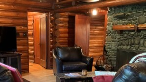 A cozy log cabin interior with a leather sofa, a small round table with a book, a leather armchair, and a stone fireplace.