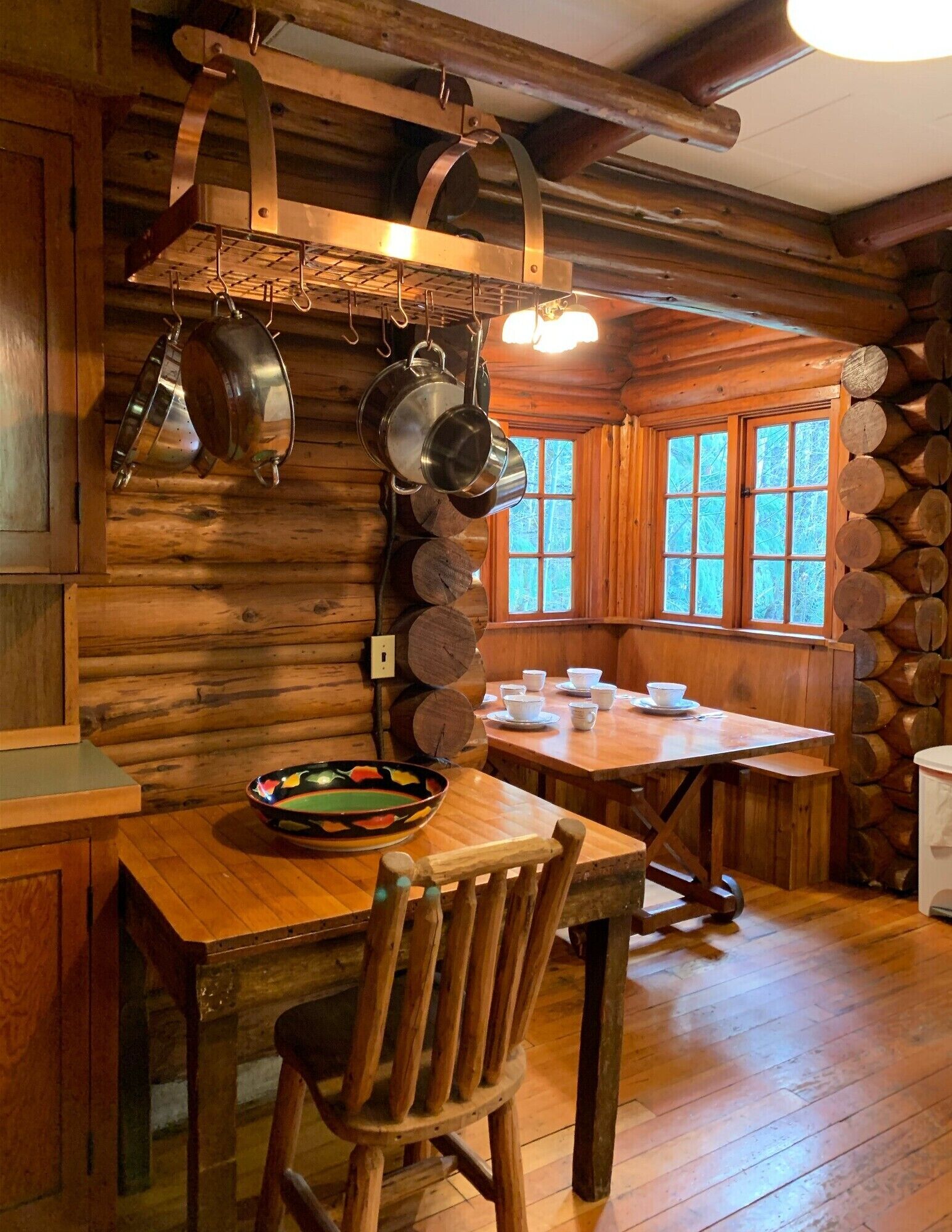 Interior rústico de una cabaña de madera con una mesa de comedor, sillas y ollas colgantes.