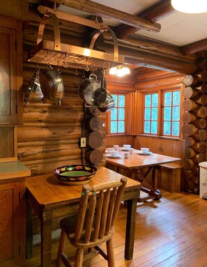 Interior rústico de una cabaña de madera con una mesa de comedor, sillas y ollas colgantes.