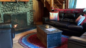 A cozy cabin interior with a stone fireplace, wooden beams, a black leather sofa, and a colorful rug.