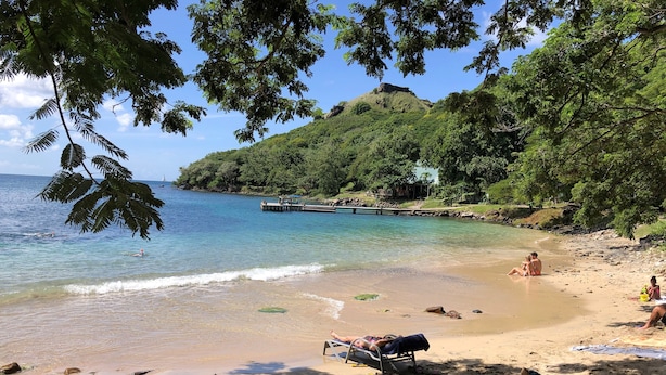 Ein Strand mit klarem Wasser, einer Anlegestelle und einer Hügelkette im Hintergrund.