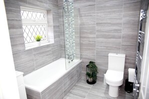 A modern bathroom with a bathtub, toilet, and a window with a plant. - Deluxe 4-bed House Close2 Wembley Stadium (London)