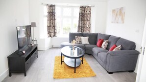 Un salon moderne avec un canapé gris, une table basse ronde, un téléviseur posé sur un meuble noir et des rideaux à motifs floraux.