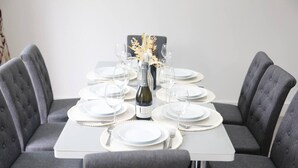 Une table à manger dressée avec de l’argenterie blanche, des verres transparents et une bouteille de vin, entourée de chaises à revêtement gris.