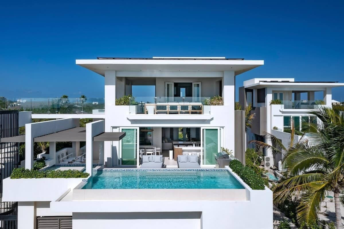 A modern two-story house with a swimming pool, outdoor seating, and a view of the ocean.