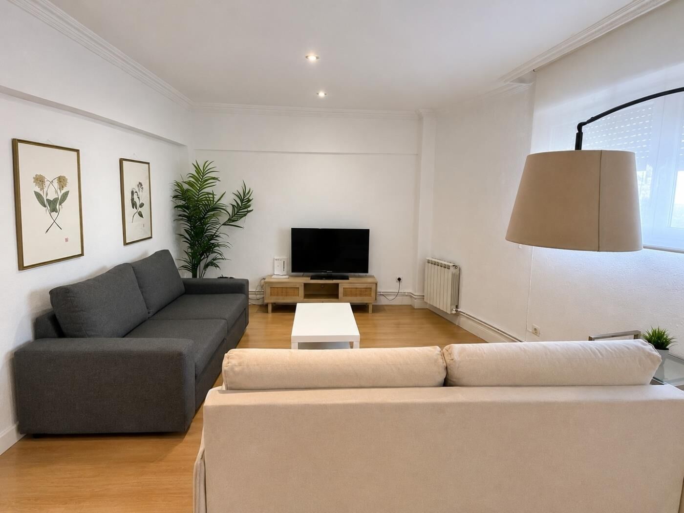 Una sala de estar moderna con un sofá gris, un sillón blanco, un televisor sobre un soporte de madera y un ventanal con una lámpara.