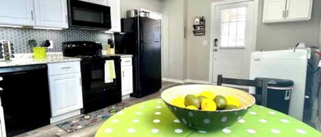 A compact kitchen with white cabinets, a black refrigerator, and a microwave. A green table with a polka dot tablecloth holds a bowl of fruit.