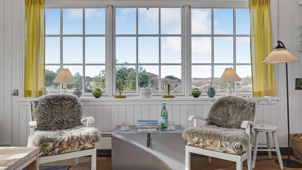 A cozy room with two white chairs with fur throws, a small table with books and a bottle, and a view of the countryside through large windows with yellow curtains.
