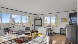 A spacious living room with a sofa, dining table, and chairs, large windows with curtains, and a fireplace.