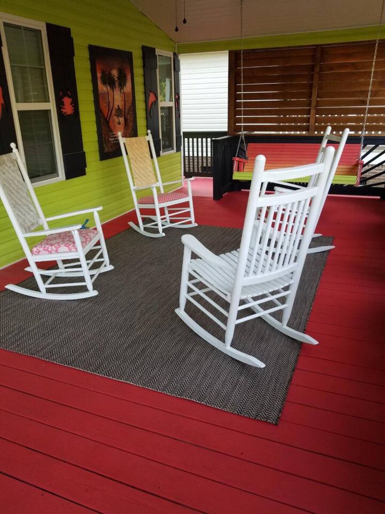 A room with green walls, a red floor, and white rocking chairs. There are paintings on the wall and a window with blinds.
