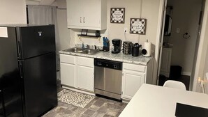 A compact kitchen with white cabinets, a black refrigerator, a dishwasher, and a countertop with various appliances and utensils.