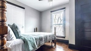 A bedroom with a bed, wooden headboard, two windows with blinds, a ceiling fan, and a dark door. - Spacious five-bedroom home in the heart of Kansas City - Fast highway access (Kansas City)