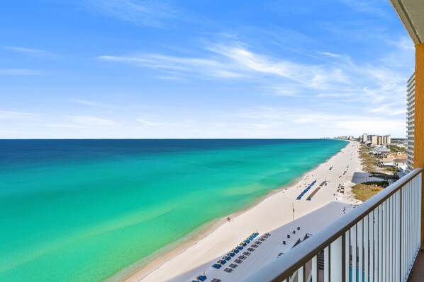A beach view from a balcony with a clear sky, a sandy beach, and a row of blue umbrellas.