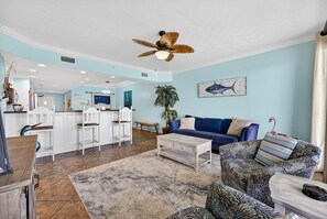A spacious living area with a ceiling fan, a blue sofa, patterned armchairs, a coffee table, and a kitchen in the background.