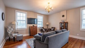 Un salon avec un canapé gris, un parquet en bois, un lustre et un téléviseur fixé au mur.