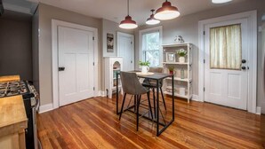 Une cuisine avec un plancher en bois, une table à manger avec des chaises, des luminaires suspendus et un meuble blanc.