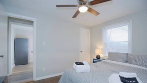 Une chambre à coucher comprenant un lit, un ventilateur de plafond, une fenêtre avec des stores et une porte donnant sur une autre pièce.