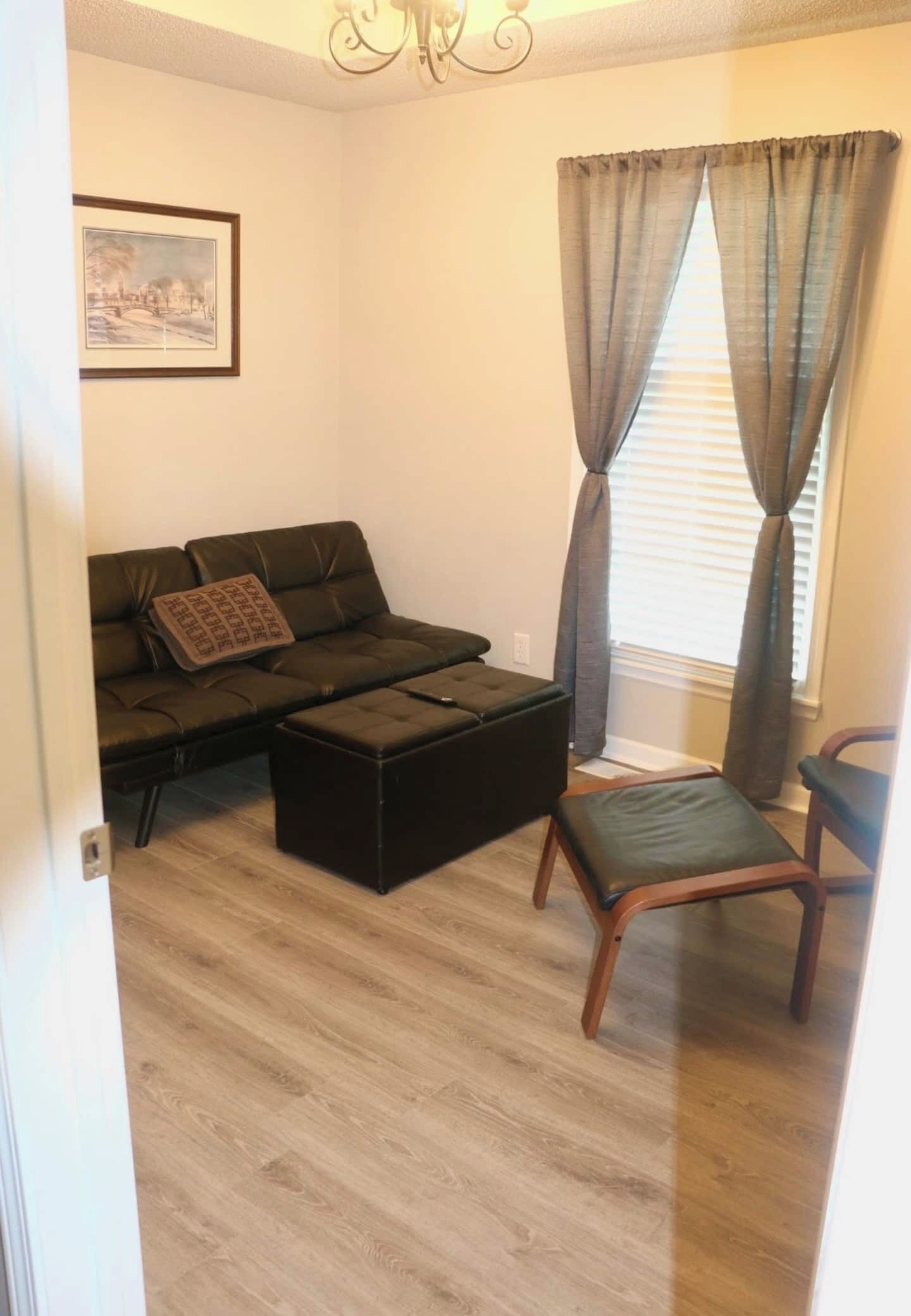 Een woonkamer met een bank, poef en fauteuil, een raam met gordijnen en een ingelijst schilderij aan de muur.