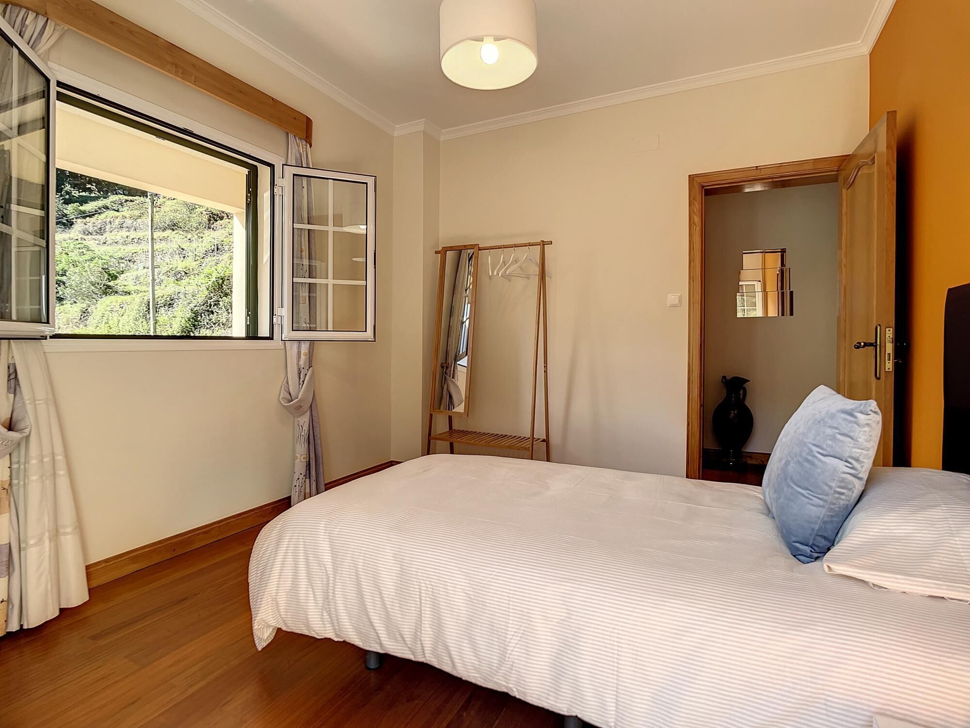 Een slaapkamer met een groot bed, een houten vloer, een raam met gordijnen, een spiegel en een kledingkast.