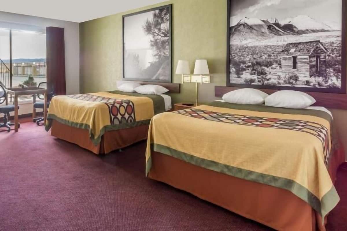 A hotel room with two beds, a desk, a chair, and a view of a building through a window.