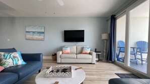 Un salon avec un canapé bleu, un canapé beige et une table basse. Un téléviseur est fixé au mur et il y a une fenêtre avec des rideaux bleus.