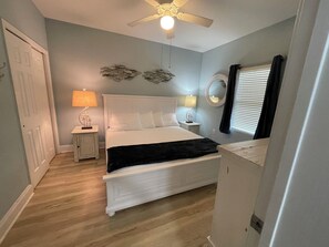 Un dormitorio con cama blanca, ventilador de techo, mesita de noche con lámpara y ventana con persianas.