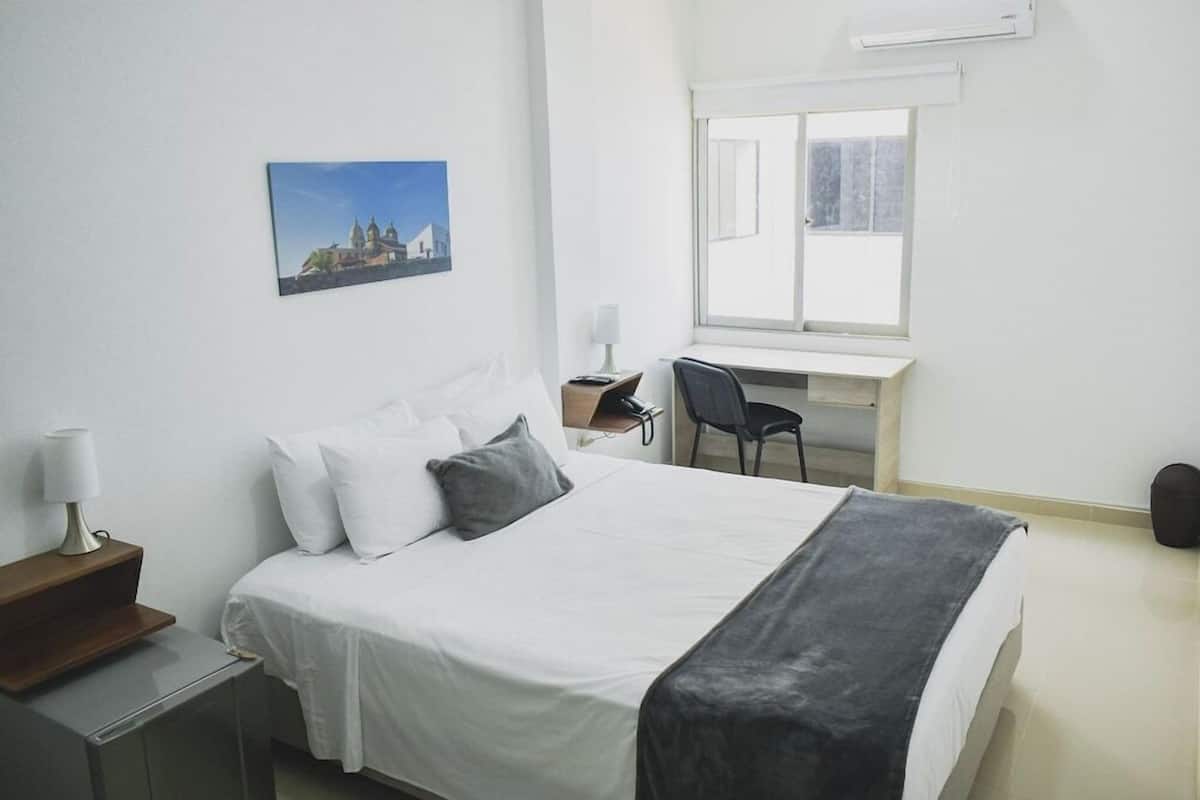 A hotel room with a bed, a desk with a chair, a window, and a picture on the wall.