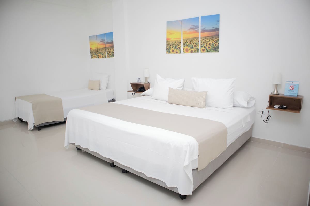 A hotel room with a large bed, a smaller bed, a bedside table with a lamp, and a wall-mounted shelf with a lamp.