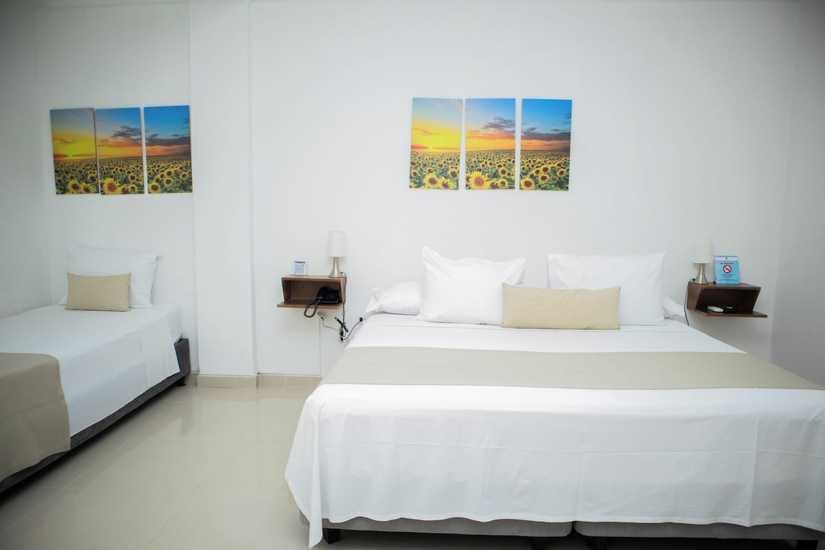 A hotel room with a large bed, two smaller beds, a nightstand with a lamp, and two wall-mounted paintings of a sunset over a field of sunflowers.
