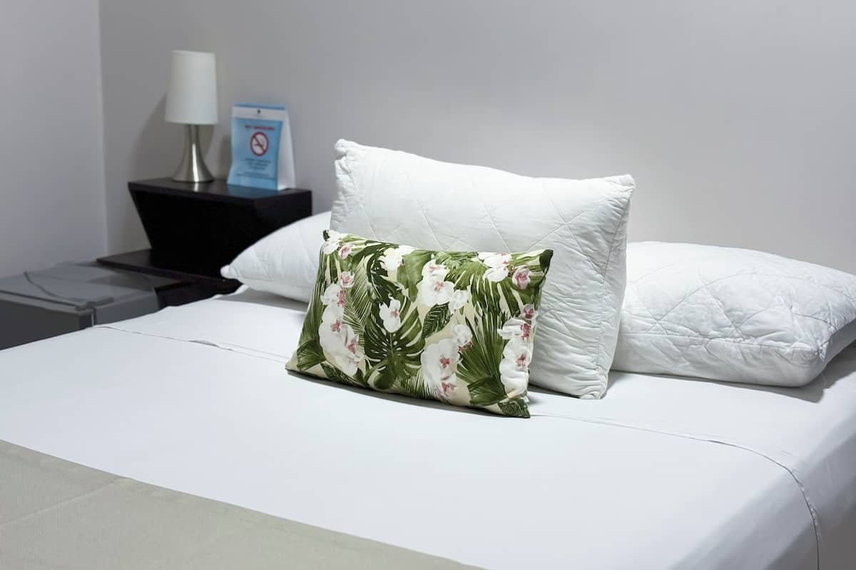 A neatly made bed with white bedding and a floral pillow. A small nightstand with a lamp and a "No Smoking" sign is visible to the left.