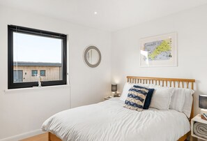 A bedroom with a large bed, a round mirror, a framed map, and a window with a view of buildings. - Poppyfields is a stunning and modern, single-storey eco lodge conveniently situated in-between Praa (Ashton)