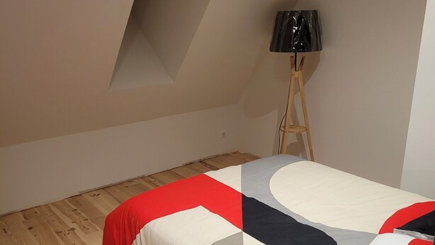 A bedroom with a sloped ceiling, a bed with a geometric-patterned bedspread, a standing lamp, and wooden flooring. - Forsythias (Messeix)