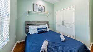 Une chambre à coucher avec un lit, une tête de lit, une armoire blanche, une étagère avec un tableau, et une fenêtre avec des stores.