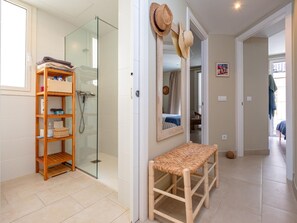 A bathroom with a glass shower enclosure, a wooden shelf with toiletries, a woven bench, and a mirror with hats hanging above it.