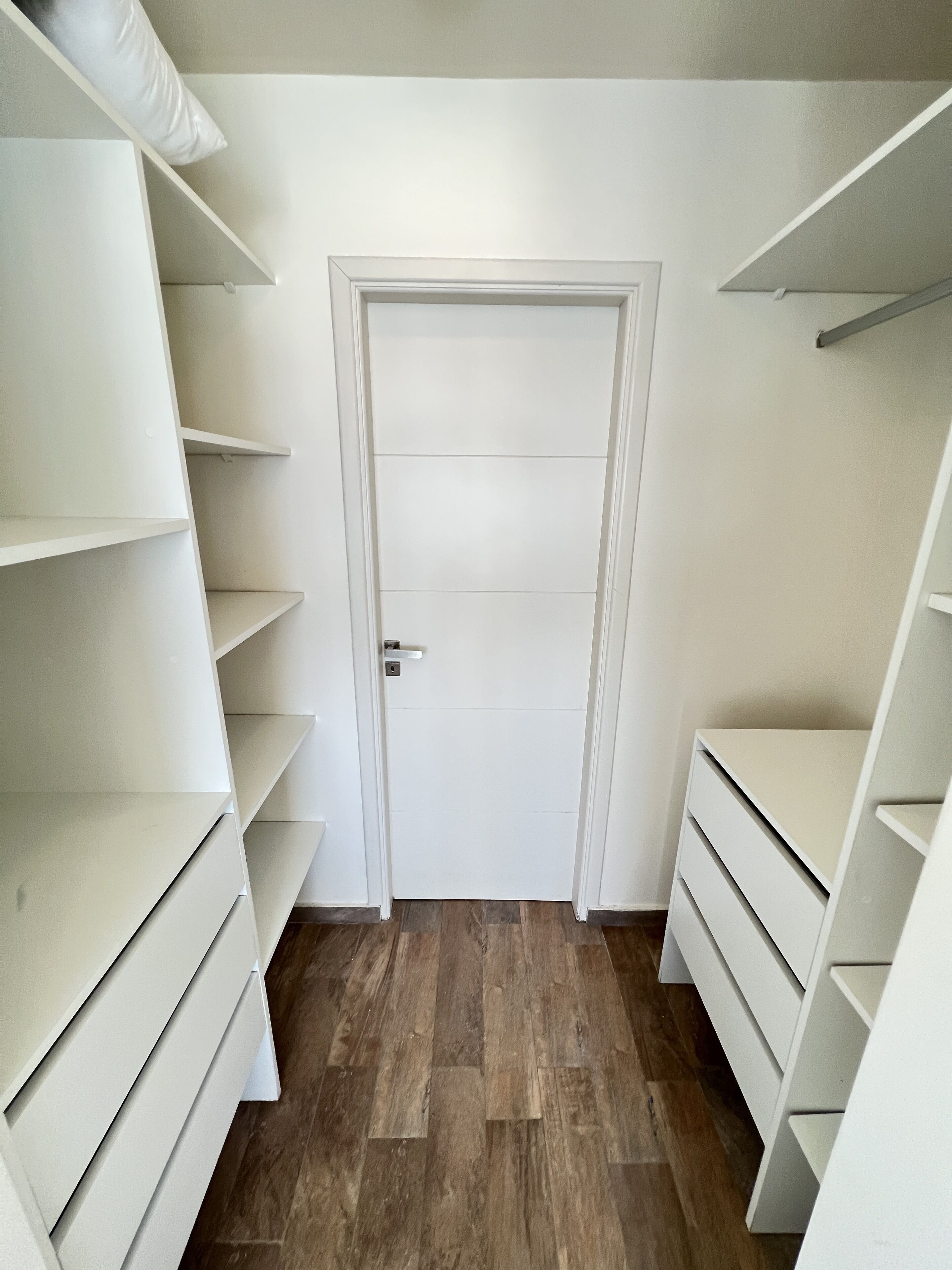 Ein begehbarer Kleiderschrank mit weißen Regalen und Schubladen, Holzboden und einer weißen Tür.
