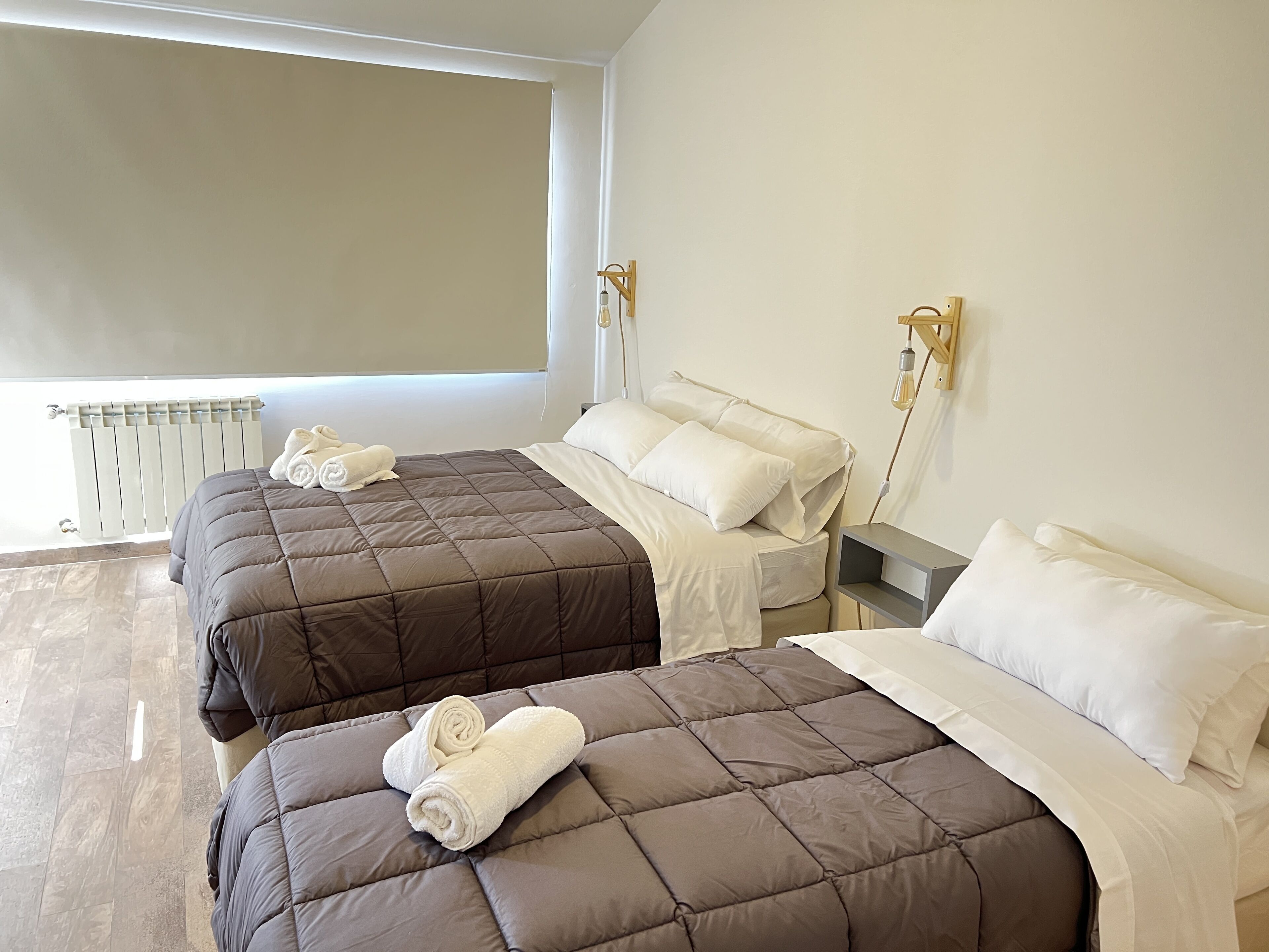 A hotel room with two beds, each with a brown quilt and white pillows. There is a window with a view of the outside, and a radiator is visible on the floor.