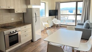 Une cuisine moderne dotée d’un four et d’un réfrigérateur encastrés, un coin repas avec une table et des chaises blanches, et une grande fenêtre offrant une vue sur la ville.